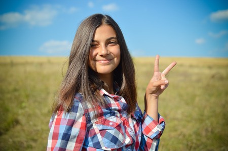 portrait of pretty teenage girl having fun showing peace sign happy smiling & looking at camera on green field under blue sky summer outdoors copy space backgroundの写真素材
