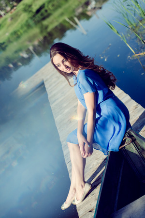 portrait image of romantic young brunette pretty lady having fun relaxing sitting in casual blue dress on river or lake pier dreaming on summer sunny day outdoors copy space backgroundの写真素材