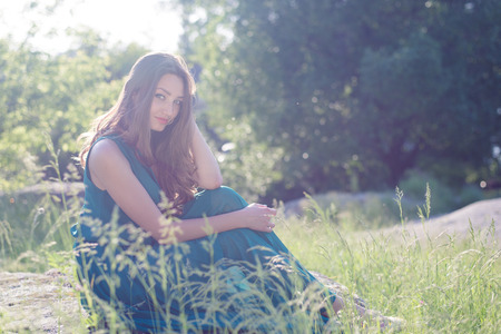 portrait of pretty fairy: beautiful young woman sitting on stone in green dress with sun light flares of rays & looking at camera happy smiling on summer outdoor copy space backgroundの写真素材