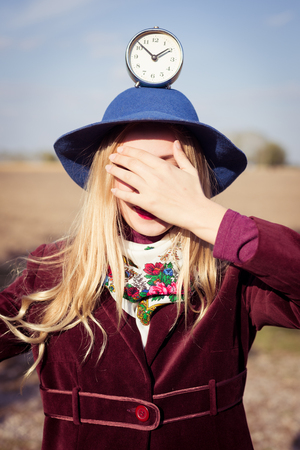 portrait of elegant beautiful blond young woman having fun holding retro alarm clock on the head eyes covered with hand on sunny autumn outdoors copy space backgroundの写真素材