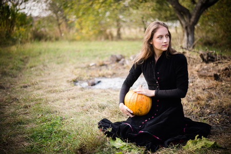 portrait of elegant beautiful blond young woman having fun holding pumpkin and looking at copy space sitting on autumn outdoors backgroundの写真素材