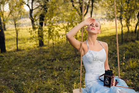 Beautiful young woman in prom or cocktail dress sitting on swings with retro photo camera on green summer or autumn outdoors copyspace backgroundの写真素材