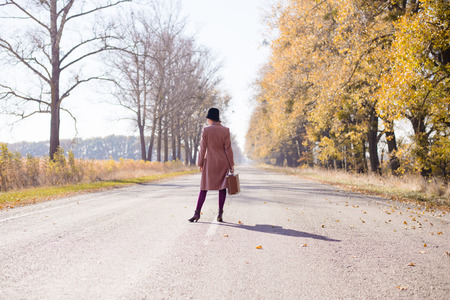 Young lady in retro hat and overcoat with vintage suitcase waiting on empty autumn roadの写真素材
