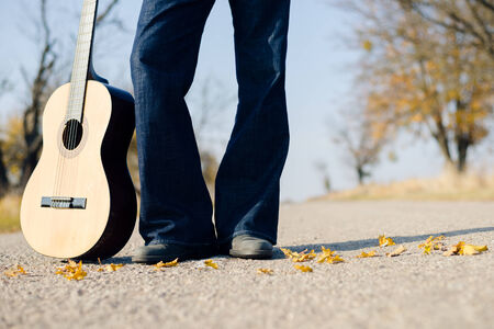 Closeup image of man musician standing with guitar in hand on autumn highway covered with yellow leaves copyspace backgroundの写真素材
