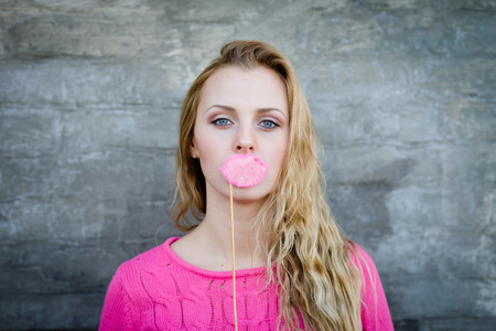 image of beautiful blond young lady in pink sweater making faces with lips props at gray concrete wall copy space backgroundの写真素材