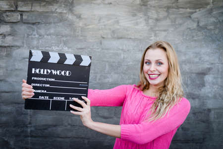 Happy young blond woman holding cinema clapper board and smiling over gray concrete or brick wall copy space backgroundの写真素材