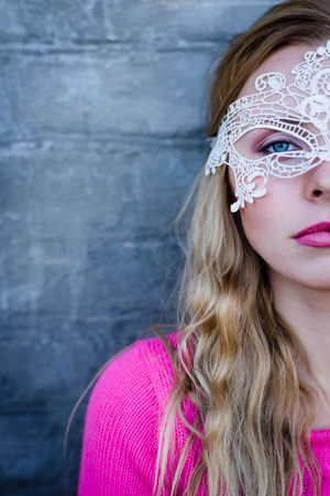 image of beautiful blond young lady in pink sweater and white lace mask half face at gray concrete wall copy space backgroundの写真素材