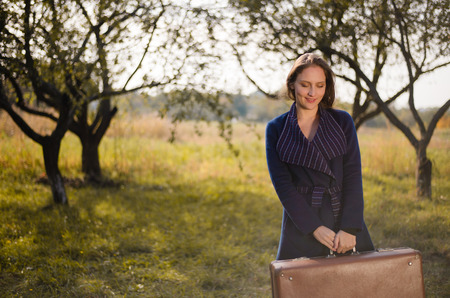 Young lady in retro overcoat with vintage suitcase waiting in autumn park copyspace backgroundの写真素材