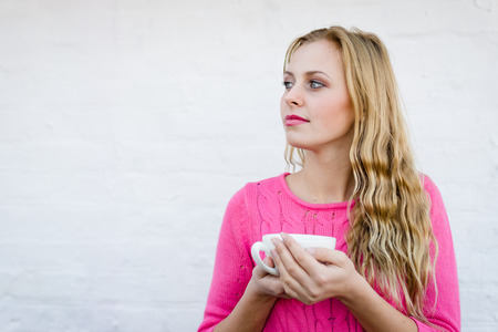 Happy young blond woman drinking hot tea or coffee and looking joyful over pin brick wall copy space backgroundの写真素材