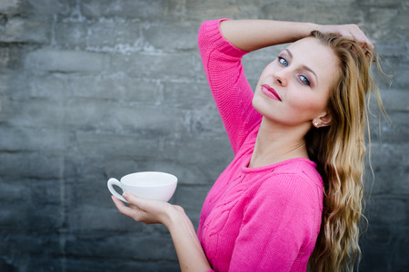 Happy young blond woman drinking hot tea or coffee and looking puzzled over gray brick wallの写真素材