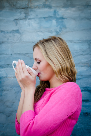 Happy young blond woman enjoying hot tea or coffee and smiling over blue brick wall copy space backgroundの写真素材