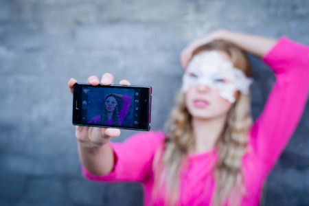 fairy selfy: image of beautiful blond young lady in pink sweater and white lace mask making selfie at gray concrete wall copy space backgroundの写真素材