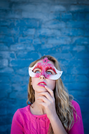 Happy young blond woman wearing carnival mask and smiling over blue brick wall with sun flare copy space backgroundの写真素材