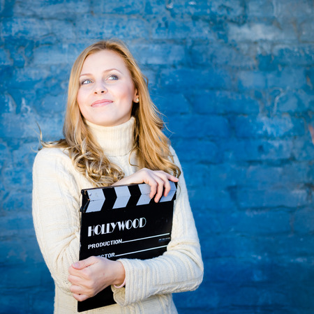 Happy young blond woman in white knitted sweater with cinema clapper board smiling over blue concrete or brick wall copy space backgroundの写真素材