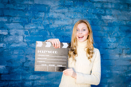 Happy young blond woman in white knitted sweater with cinema clapper board smiling over blue concrete or brick wall copy space backgroundの写真素材