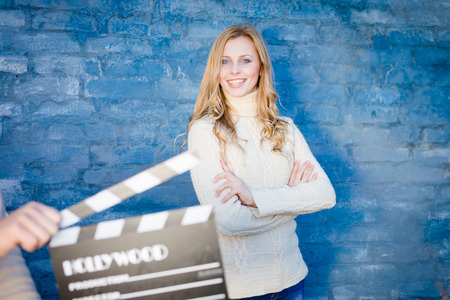 Happy young blond woman in white knitted sweater with cinema clapper board smiling over blue...