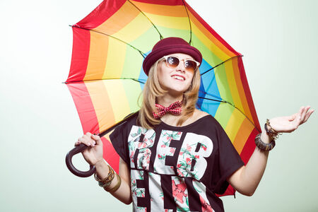 Studio portrait of teenage hipster girl wearing trendy hat and sunglasses with colorful umbrella...