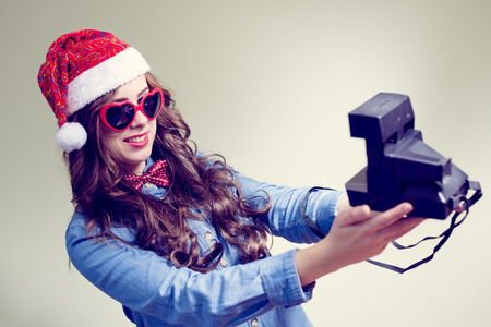 Studio portrait of funny hipster girl wearing Christmas hat and heart shape sunglasses making selfie with retro photo camera over pale green copy space backgroundの写真素材