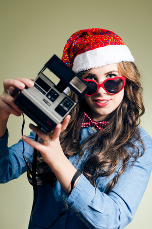 happy smiling & holding retro photo camera studio portrait of funny hipster girl wearing Christmas hat and heart shape sunglasses over pale green copy space backgroundの写真素材