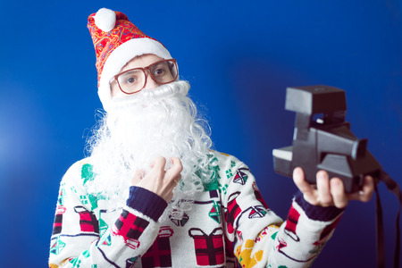 studio portrait of funny young hipster man in santa beard making selfi with retro camera on blue backgroundの写真素材