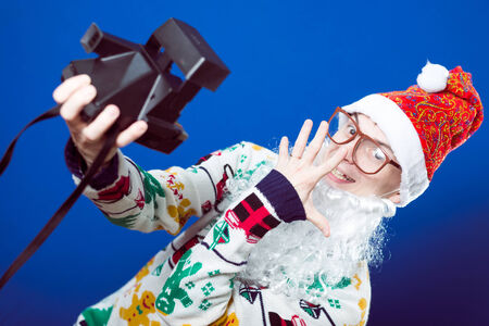 Funny young hipster man in Santa beard making selfie with retro camera on blue background studio portraitの写真素材