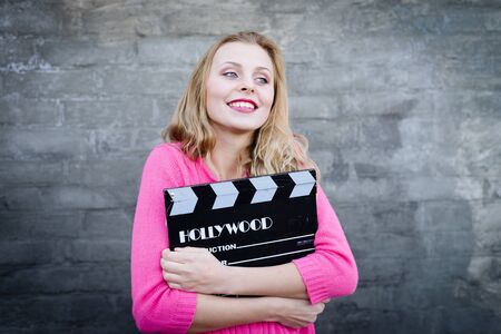 Happy young blond woman in pink knitted sweater with cinema clapper board smiling over brick wall copy space backgroundの写真素材