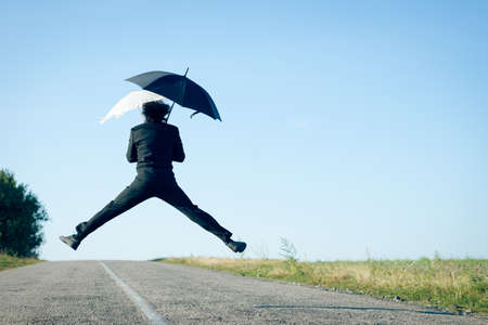 Rear view of man jumping with two umbrellas on the roadの写真素材