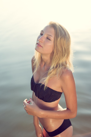 Young beautiful blond woman with eyes closed wearing black bikini relaxing in water on summer day outdoors backgroundの写真素材