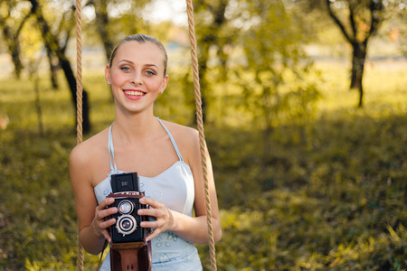 Beautiful young woman in prom dress sitting on swing with retro photo camera on green summer...
