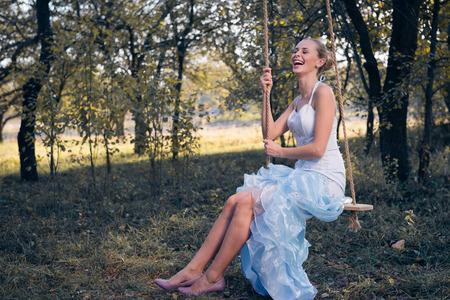 Beautiful young woman in prom dress sitting on swing on green summer outdoorsの写真素材