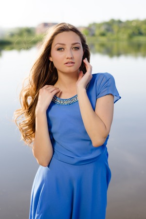 Half-portrait of sensual beautiful brunette female with long hair in blue dress standing by lakeの写真素材