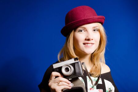 Close up picture of young smiling lady in red hat taking picture with retro camera over deep blue...