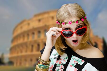 Portrait of excited woman in flower wreath and heart shaped glasses. Fancy girl shocked on Coliseum blurred background.の写真素材