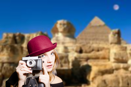 Picture of joyfull woman in hipster glasses and pot hat with vintage camera. Young traveler smiling on Egypt pyramid blurred background.の写真素材