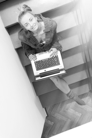 black and white image of blond young pretty businesswoman or student having fun working typing on laptop computer relaxing sitting on stairs at home, happy smiling and looking up at camera portraitの写真素材