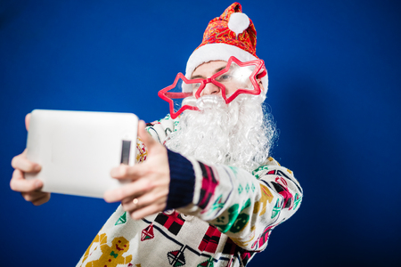 Funny hipster Santa Claus wearing super size star shape eyeglasses and cute gingerbread sweater on blue background taking selfie with tablet pcの写真素材