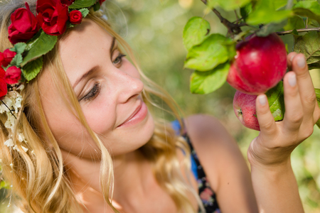 Closeup of young beautiful lady smiling among red apples with wreath of flowers. Blond sexy girl on summer outdoor copy space backgroundの写真素材