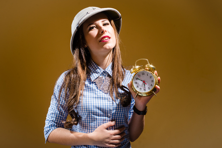 Fuuny young woman in checkered shirt and pith helmet holding alarm clock over brown backgroundの写真素材