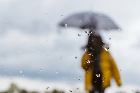 Blurred picture of woman standing under umbrella with raindrops on window glass foreground under...