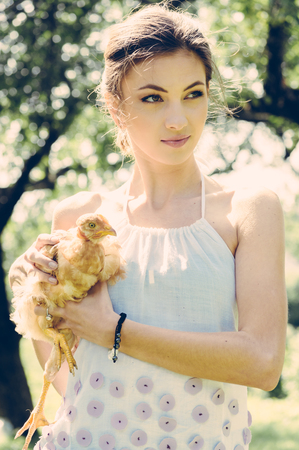 Beautiful happy smiling young woman holding a live little red chicken in nature summer backgroundの写真素材