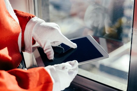 Santa Claus standing at window and using apps on a touch screen tablet in white gloves, Christmas and technology conceptの写真素材
