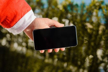 Santa Claus holding black modern model of mobile smartphone ready for Christmas time on green pine backgroundの写真素材