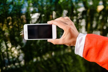 Santa Claus holding white modern model of mobile smartphone ready for Christmas time on emerald pine backgroundの写真素材