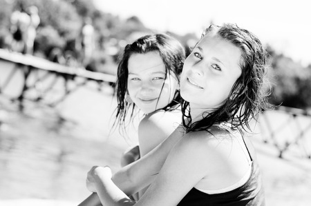 Black and white image of 2 charming girl friends sitting near lake looking at camera. having fun relaxing on the river bank sandy beach ground, summer outdoors background, close up portraitの写真素材