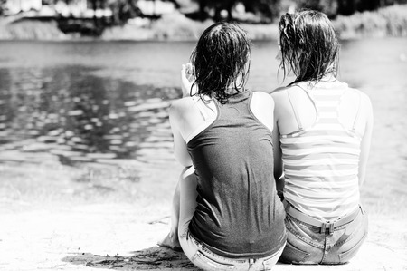 Black and white image of 2 charming girl friends sitting near water. having fun relaxing on the river bank sandy beach ground, summer outdoors background, back viewの写真素材