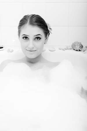 Black and white image of attractive young brunette woman having fun relaxing taking a bath foam surrounded with sea shell orchid flower gently smilingの写真素材
