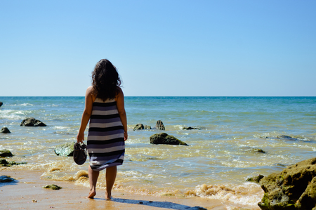 Back view of elegant young lady enjoying summer beach and sun, standing in water. Happy tired traveler contempatingの写真素材