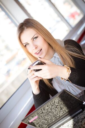 Pretty woman with hands holding coffee cup and mobile phone on restaurant background, mockup space for text message or design. closeup portrait viewの写真素材