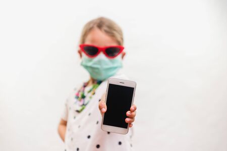 Little blond girl wearing disposable mask and sunglasses holding mobile phone over white backgroundの写真素材