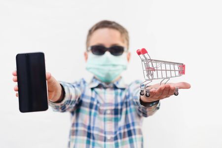 Little school boy wearing disposable mask and sunglasses and holding small shopping cart and mobile phone over white backgroundの写真素材